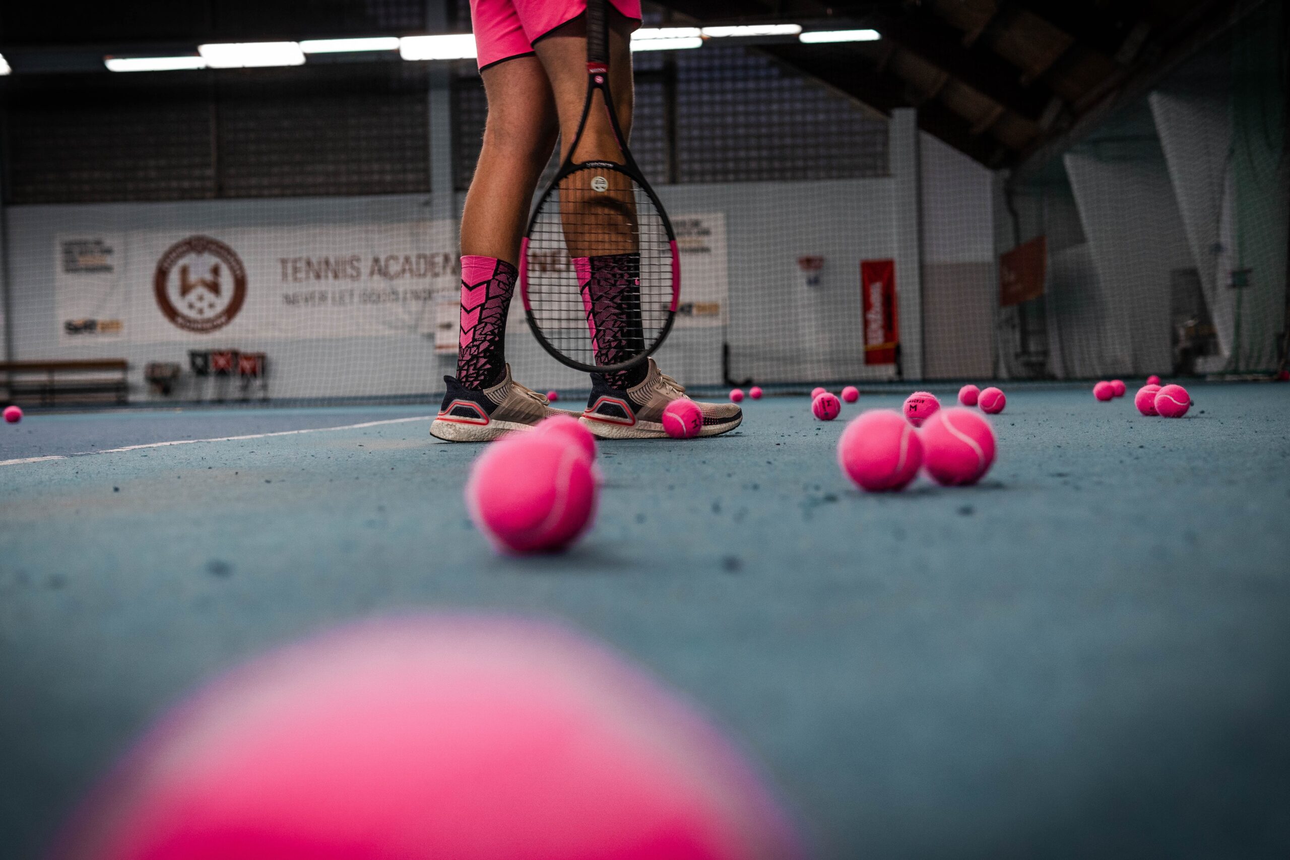Tennis player with pink uniform, pink tennis balls lying around the court. Sign in the background reads "Tennis Academy"